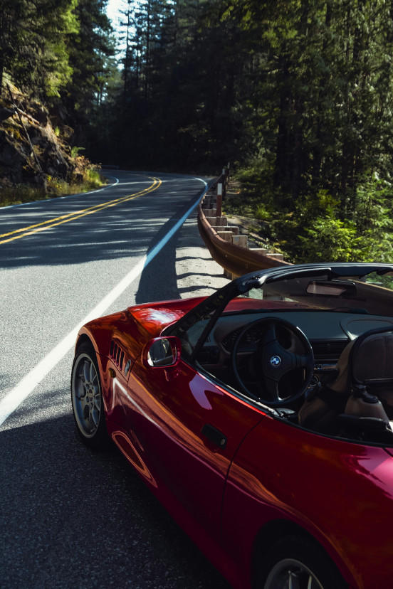 A red convertible parked at a small pull-off on the right, alongside a curving asphalt road lined with coniferous trees.